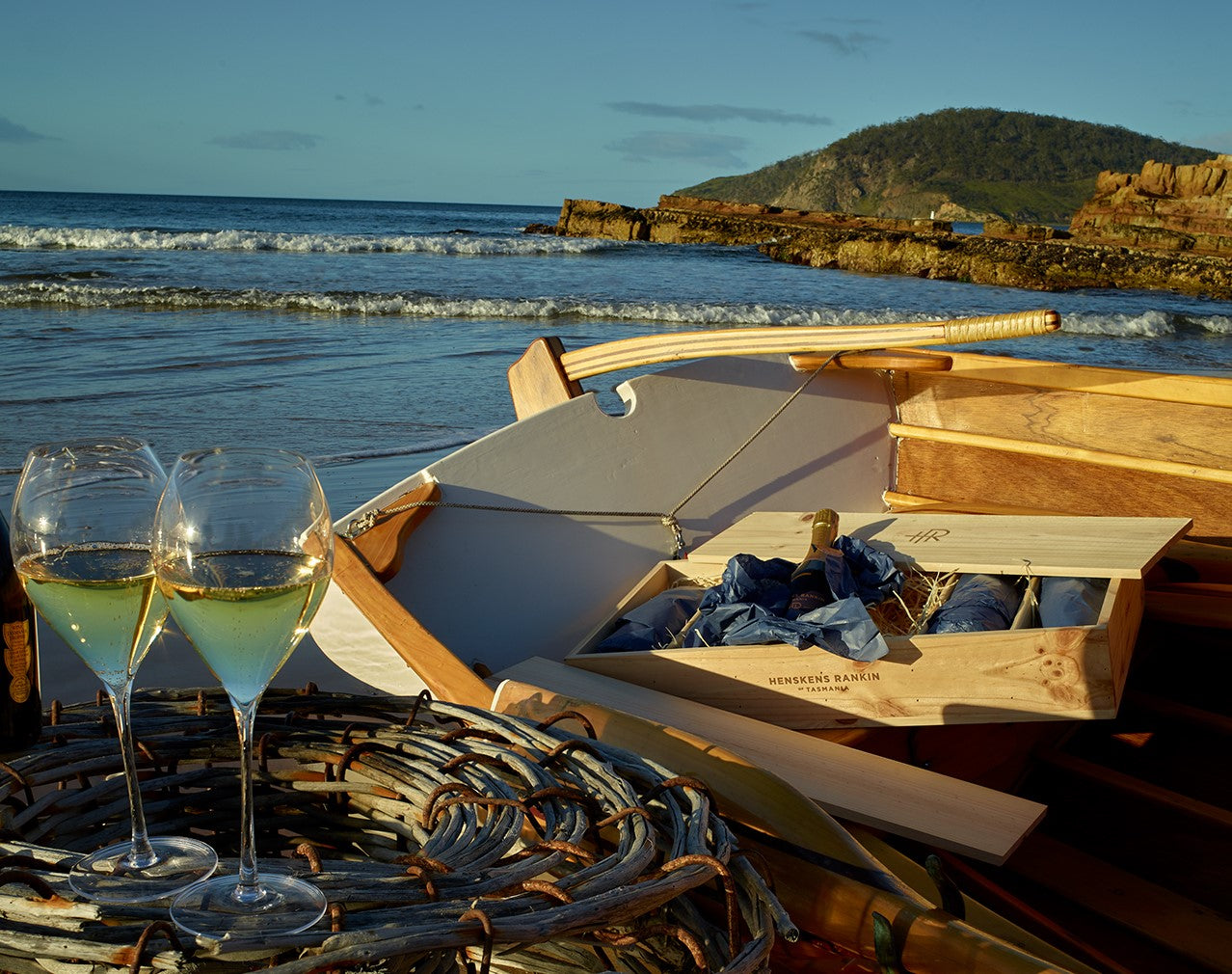 Wine glasses perched on a crayfish pot next to a boat with a crate of wine inside on a beach